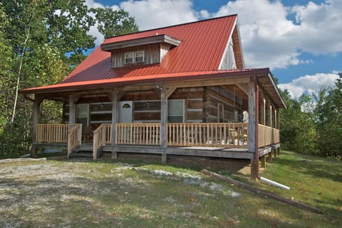 Exterior view of the log cabin with a red metal roof, wraparound porch, and wooden railings surrounded by trees.