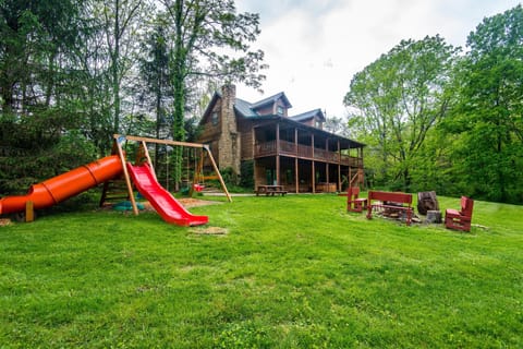 Outdoor play area with red slides and swings beside a wooden lodge and a fire pit with red benches.