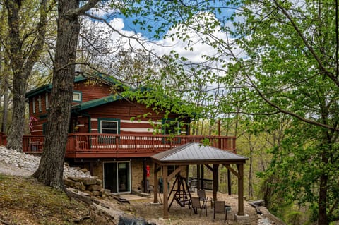 Exterior view of the log cabin showing red wood siding, green-trimmed windows, wraparound deck, and covered patio with seating.
