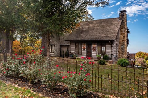Front view of a log cabin with stone chimney, wooden siding, autumn wreaths by the door, and a red rose garden fenced in front.