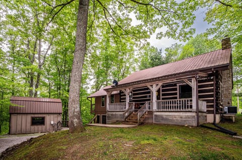 Exterior view of a rustic log cabin with a covered porch, stone chimney, and a detached brown shed surrounded by trees.