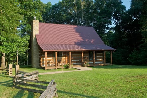 Front view of the log cabin with a red metal roof, stone chimney, wraparound porch, and lawn bordered by rustic wooden fencing.