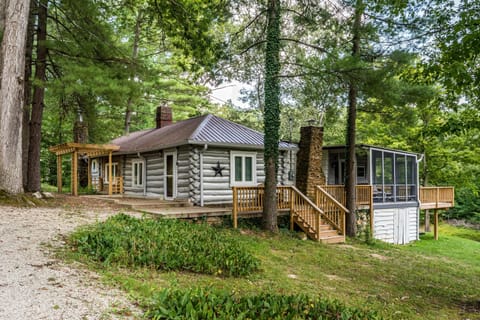 Exterior view of the 1930s log cabin with screened porch, stone chimneys, wooden deck, and surrounding trees.