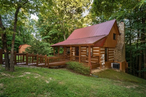 The log cabin exterior features a red metal roof, stone chimney, and wraparound wooden deck surrounded by lush trees.
