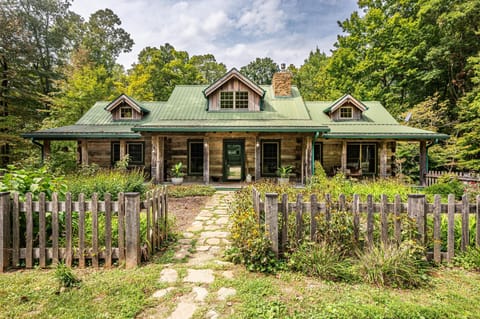Stone pathway leads through a weathered wooden fence to a log cabin with green metal roof and stone chimney.