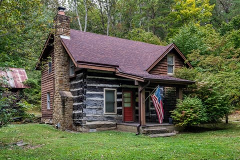 Front view of a log cabin with a stone chimney, red door, covered porch, and an American flag amidst green trees.