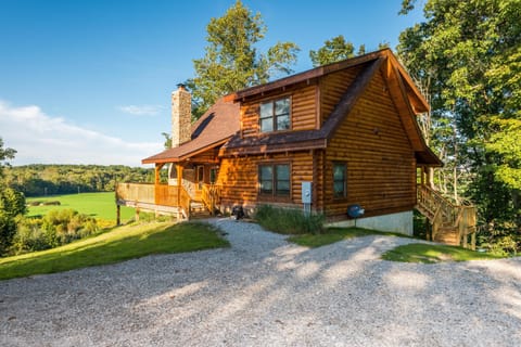 Log cabin exterior with gravel driveway, stone chimney, wraparound wooden deck, and expansive green fields beyond.