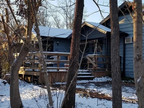 Snow-dusted wooden deck wraps around the blue cottage, framed by leafless trees under clear winter skies.