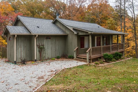 Exterior view of green wood-paneled cottage with covered porch, gravel driveway, and autumn trees surrounding.