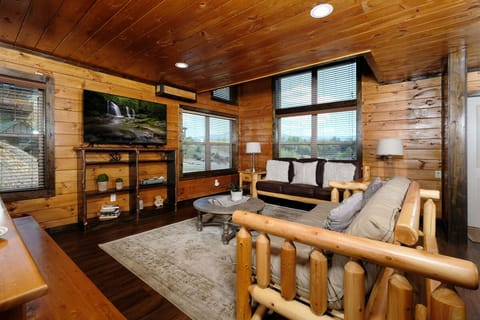 Cozy log cabin living area featuring rustic wood furniture, large windows with blinds, and a TV showing a waterfall.