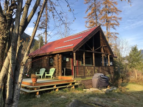 Exterior view of a log cabin with a red metal roof, covered porch, wooden deck, and light green Adirondack chairs.
