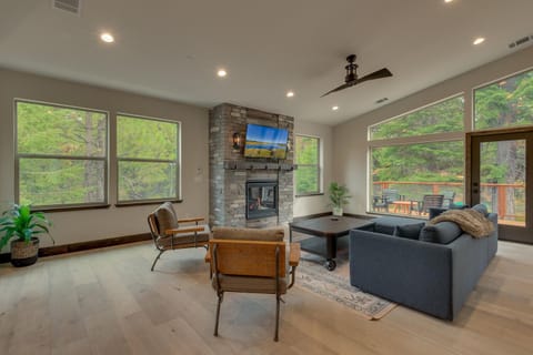 - Bright living room with forest views all around  
- Stone fireplace centerpiece and modern furnishings  
- Direct access to outdoor deck and fresh mountain air