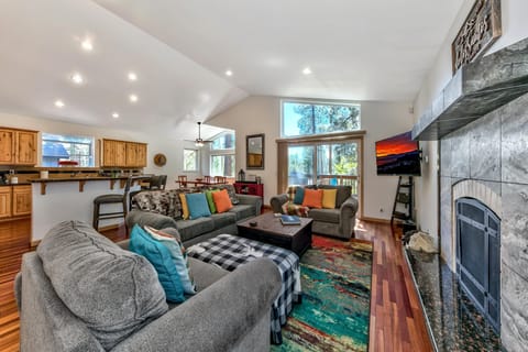 A living room with gray sofas, colorful pillows, a patterned rug, and a stone-tiled fireplace with a mounted TV.