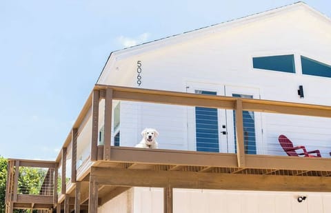 This photo captures a bright, airy scene of a white beach-style house or elevated cottage under a clear, pale blue sky. The composition is angled from a low perspective, looking up at the second-story deck.