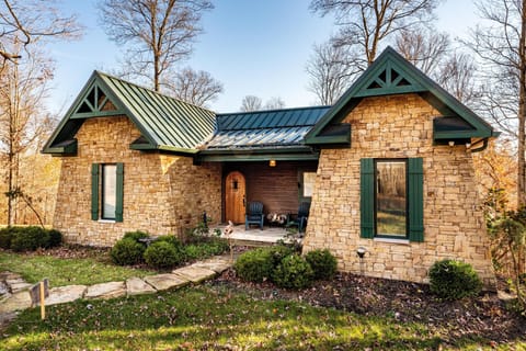 Stone exterior with green-trimmed windows and metal roof, leading to a small covered porch with two chairs.