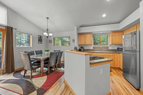 Spacious open-concept dining area with elegant chandelier lighting and a fully equipped kitchen featuring natural wood cabinetry and abundant counter space. Large windows provide plenty of natural light and a tranquil wooded view.