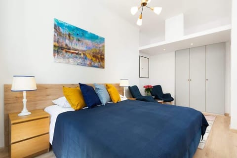 A cozy bedroom with a blue bedspread and decorative yellow pillows. A bedside table with a lamp adds a touch of warmth to the space.