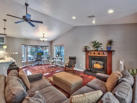 Living room with plush sectional, patterned ottoman, wood-trimmed gas fireplace, and dual desks near bay windows.