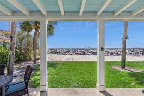 Covered patio with beachfront access, featuring lush lawn, palm trees, and a rocky shoreline framing calm coastal waters—ideal for morning coffee or sunset relaxation.