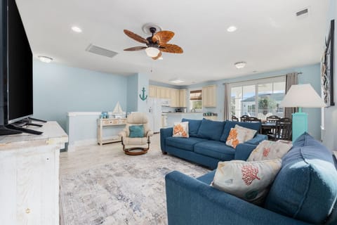 The open-concept living area features cozy blue sofas with playful coral-patterned pillows, creating a relaxed coastal vibe. Large windows and sliding glass doors let in plenty of natural light and offer easy access to the sunny balcony.