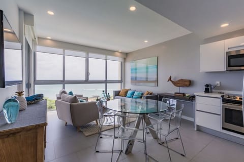 Your open-concept living space flows seamlessly, with a sleek kitchenette and dining area opening to a cozy lounge—perfect for gathering after beach days, with calming sea views beyond.