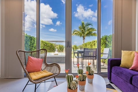 Curl up in your stylish living room as sunlight streams through floor-to-ceiling glass doors, framing vibrant tropical greenery and distant water views beyond your private patio.