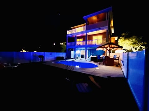 Marine Life OBX backyard lit up at night, outdoor tv, tiki bar, pool, hot tub, gas grill, turf grass, fenced