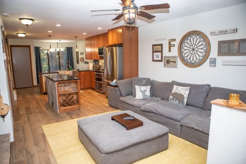 Open-concept living room and kitchen with wood flooring, gray sectional sofa, a central ottoman, kitchen island with wine storage, stainless steel appliances, and wall-mounted decorations.