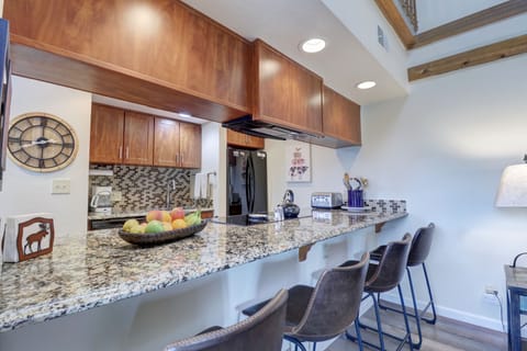 Modern kitchen with granite countertops, wooden cabinets, mosaic backsplash, a bowl of fruit, black fridge, and three bar stools. Lighted clock on wall and decorative wall art visible.