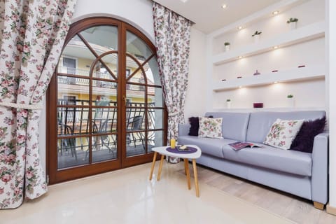 A spacious living room featuring a cozy sofa, a coffee table, and modern wall-mounted shelves illuminated by spotlights. The large window lets in natural light, highlighting the neutral tones.