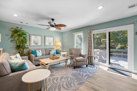 A bright, coastal-inspired living room featuring seafoam walls, beach-themed décor, and sliding glass doors that open to a spacious deck surrounded by lush greenery.