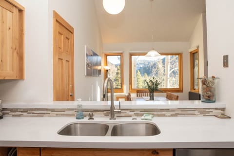 A modern kitchen with a double sink and white countertops. In the background, a well-lit dining area features large windows with a view of trees and mountains. A wooden door and cabinets are on the left.