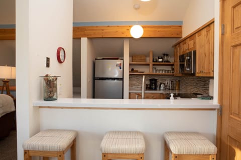 A small kitchen area with a counter bar, three wooden stools, a fridge, microwave, and storage shelves in the background. A round light hangs from the ceiling. There's a clock on the wall.