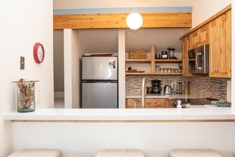 A small kitchen features wooden cabinetry, an overhead light, a clock on the wall, a silver fridge, various kitchen appliances, and a white countertop with stools in the foreground.