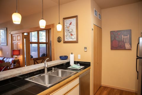 A kitchen interior with a double sink, black countertop, and stainless steel dishwasher. Two framed pictures are on the beige walls, and three pendant lights hang from the ceiling.