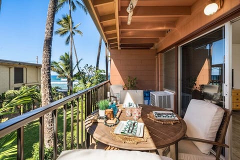Covered balcony with wicker dining set, soft cushions, and peaceful tree‑top views framed by warm island sunlight.