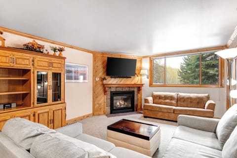 Snow Flower 43's living room has a large window framing a mountain and forest, grey sectional, leather couch, and fireplace.