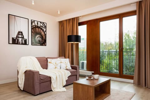 A cozy living room with a sofa, a coffee table, and a TV mounted on the wall. Large windows with wooden frames provide plenty of natural light and offer a view of the greenery outside.