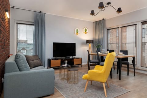 A modern living room featuring a comfortable sofa, a large flat-screen TV, a coffee table, and a yellow armchair, with large curtains covering the windows.