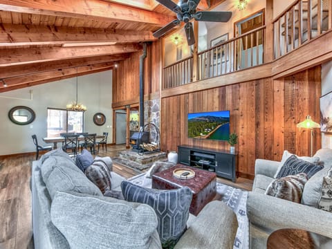 Living room with vaulted wood ceilings, stone fireplace, gray sectional sofa, and wall-mounted TV above black console.