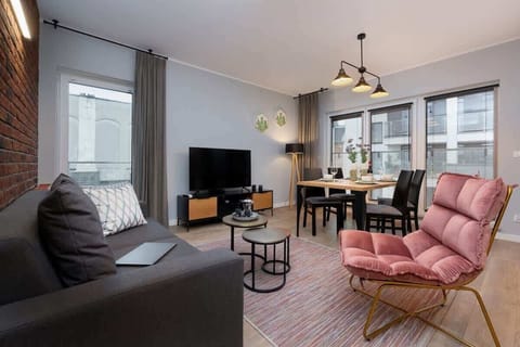 A stylish living room with light-colored walls, gray sofa and pink chair, a coffee table, a large window lettin in natural light, and a modern chandelier.