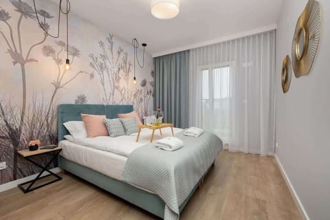 A modern bedroom with a large bed, blue accents, floral wallpaper, and sheer curtains allowing natural light.