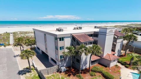 Three-story condo building with light stucco walls, red tile roof accents, and palm trees by a sandy beach.
