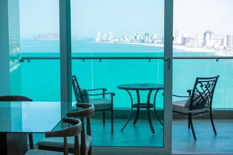 Glass sliding doors open to a balcony with wrought iron chairs and a round table overlooking the ocean and city skyline.