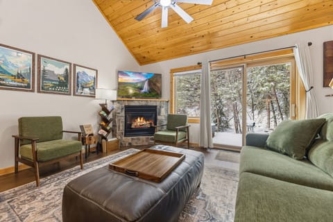 Unwind in your cozy living room with crackling fireplace, plush green seating, and snowy forest views through sliding glass doors.