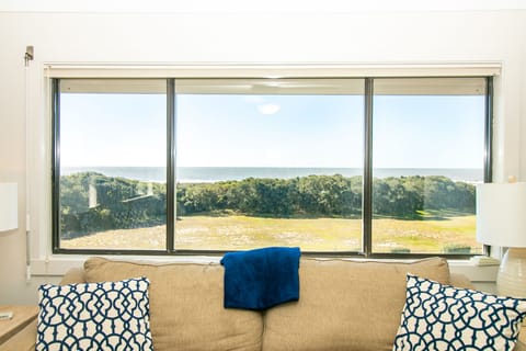 Bright living room with beige sofa, patterned pillows, coffee table, side lamps, large ocean-view window, sliding glass door, carpet, and board games.
