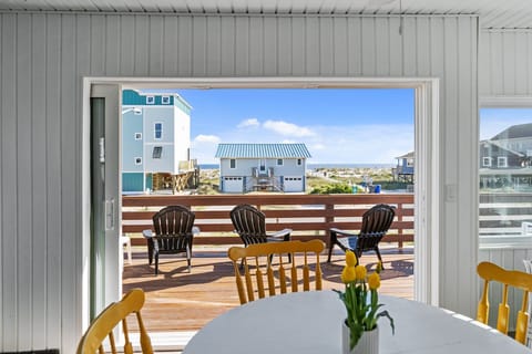 Dining area with a round white table, yellow chairs, sliding doors, deck seating, and a bright ocean view beyond neighboring beach homes.