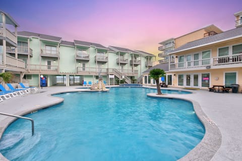 Beachside resort complex featuring a sparkling pool courtyard surrounded by colorful multi-story accommodations under vibrant twilight skies.
