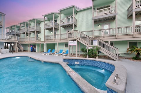 Multi-story beachside property featuring mint green units with private balconies, shared pool and hot tub area during twilight hours.
