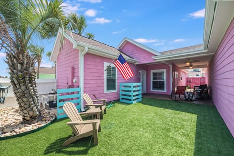 Colorful coastal cottage featuring a charming pink exterior with lush artificial turf lawn, palm trees, and comfortable outdoor seating for relaxation.